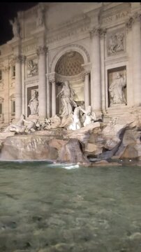 A vertical video pan from top to bottom of the magnificent Trevi Fountain in Rome, Italy, showcasing the Baroque sculptures of Oceanus, the Tritons, and the wild horses, and the turquoise water.
