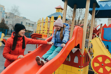 A happy, smiling teenage girl slides down a playground slide outdoors with her caring young mother. Photo, childhood concept.