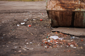 Obraz premium Litter scattered on dirt ground near rusty metal dumpster outdoors. Urban waste scene with cups and debris. Pollution, neglect, environment, cleanup concepts.