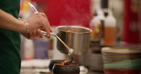Ladling soup. Pouring food. Chef measures portion of hot meal from metal pan to small cauldron. Professional cookware for kitchen oven.