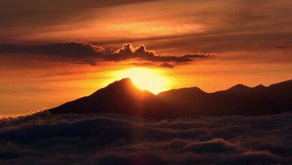 Bali, Mount Batur volcano silhouette rising above a layer of soft clouds during a vibrant golden and orange sunset, creating a serene and breathtaking natural landscape. Aerial view panorama