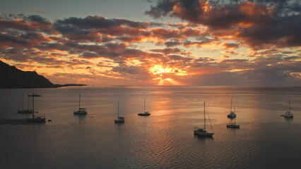 Moorea island landscape showcasing an aerial view of anchored sailboats on the calm ocean water, reflecting the warm colors of a dramatic sunset sky with sun rays breaking through colorful clouds © Goinyk