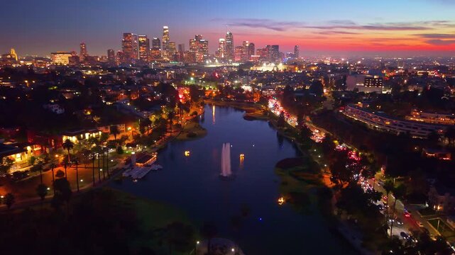 A dramatic aerial view at night captures the brilliant, illuminated skyline of downtown Los Angeles reflecting across the dark, placid surface of Echo Park Lake