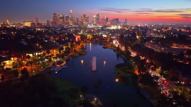 A dramatic aerial view at night captures the brilliant, illuminated skyline of downtown Los Angeles reflecting across the dark, placid surface of Echo Park Lake