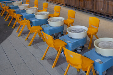 Empty children's pottery workshop with row of small electric pottery wheels (blue bases, white wheel heads) paired with bright yellow plastic chairs, ready for kids' ceramics class.