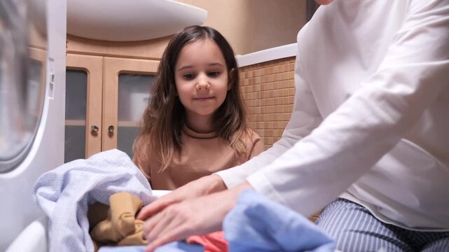 Mother and daughter share a happy moment doing laundry together by a front loading washer, folding clothes from a full basket and learning household chores at home