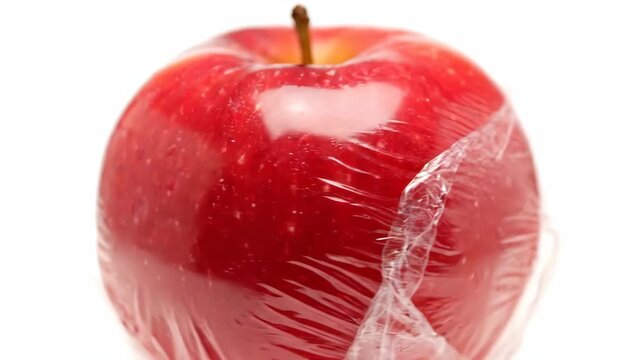 Fresh red apple in plastic wrap slowly turning. Food preservation, hygiene, and environmental concerns about single-use plastics