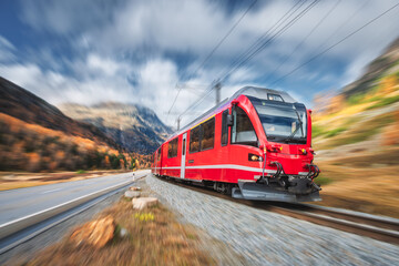 Red modern passenger train is moving on railway in swiss alps at sunset in autumn. High speed train in mountains, road, orange trees in fall with motion blur effect. Bernina Express, Switzerland