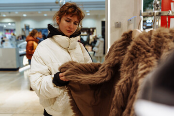 portrait of red-haired attractive young woman trying on fashionable fur coat in store