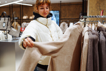 portrait of red-haired attractive young woman trying on fashionable fur coat in store