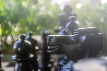 A black chess Knight piece in close-up detail, with other blurred black pieces and a green foliage background. © Afi Jamal