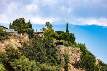Obraz premium Ottoman-era house with tile roof and chimney perched on a lush rocky cliff among cypress trees under a classic Mediterranean blue sky. Kaleiçi, Antalya, Turkey, Mediterranean.