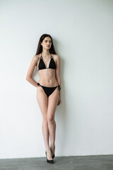 Fototapeta premium Slim fit young woman in black bikini posing in studio, healthy athletic body, minimal swimwear fashion, confident female model, beauty and fitness concept, modern clean background photography