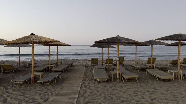Beach in Chania, Greece, at dusk with straw parasols and empty sun loungers facing the calm sea. Coastal scene with pastel sky and evening light over the horizon.