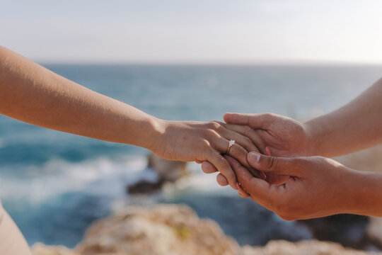 Romantic close-up of a couple holding hands with a diamond engagement ring on the woman's finger against a scenic rocky coastline and blue sea