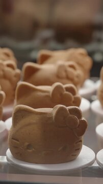 Close-up of cute cartoon cat-shaped sponge cakes on white trays in a bakery display, showing rows of golden-brown molded pastries with bows and whisker details under lighting