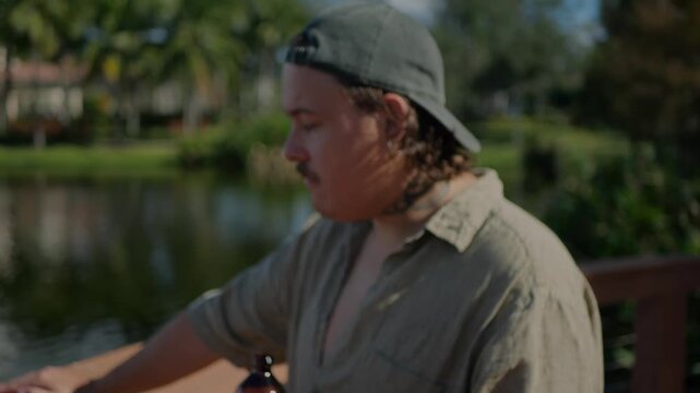 Tattooed young man in casual shirt and backwards cap drinking from dark glass coffee bottle outdoors by lake, enjoying refreshing beverage in warm sunlight, lifestyle scene