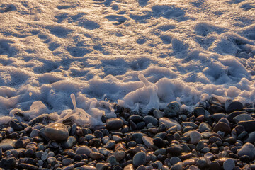 Golden sunset light illuminates the white sea foam cascading onto the wet pebbly shore. Creating a beautiful natural detail of the wave washing ashore. Great for nature concepts.