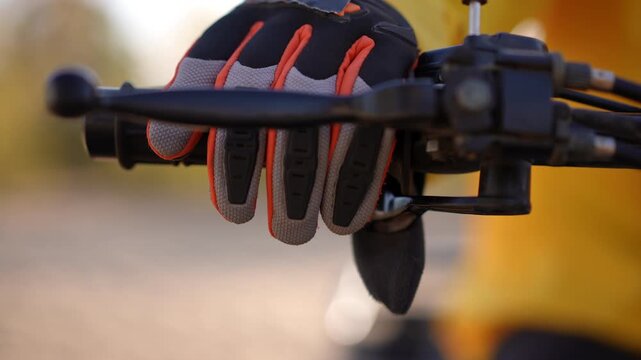 Gloved hand on motorcycle throttle, closeup view of rider adjusting grip and brake lever, orange accented protective glove and yellow jacket sleeve, shallow