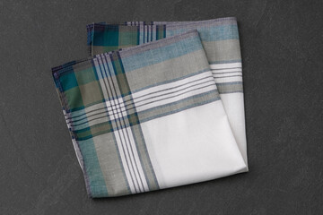 A neatly folded white handkerchief with a green, teal, and gray plaid border on a dark gray, textured surface.