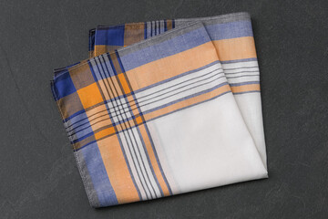 A neatly folded white handkerchief with a yellow, blue, and black plaid border on a dark gray, textured surface.