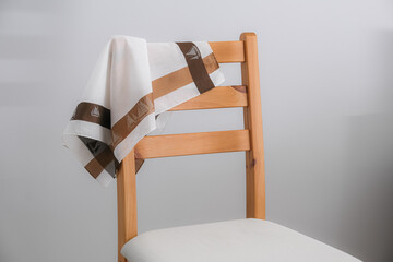 A white cotton handkerchief with a wide brown and tan border, featuring a sailboat pattern, is casually draped over the back of a light wooden chair with a white padded seat.