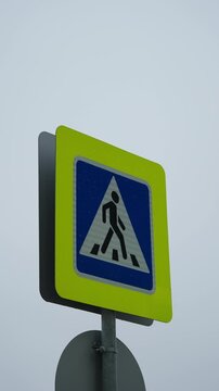 Close-up vertical view of a high-visibility pedestrian crossing traffic sign with fluorescent yellow border and blue triangle, mounted on a pole against an overcast sky to highlight urban road safety