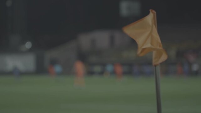 Close up of orange corner flag waving in wind during night soccer match. Background features blurred players and stadium lights with cinematic shallow depth of field effect.

