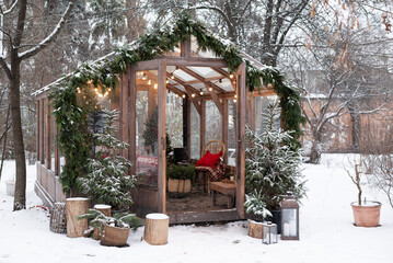 Cozy wooden winter greenhouse decorated for Christmas with lights, garlands, and small snow-covered pine trees in the yard.