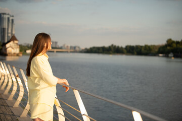 Fototapeta premium A woman stands by a white railing with a river in front. She looks out at the water and city buildings in the background. The scene is in the late afternoon