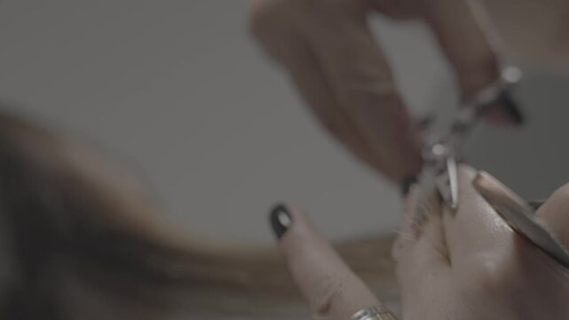 Close-up view of professional hairdresser hands with black nails carefully trimming damaged split ends of red hair using comb and sharp scissors in beauty salon.

