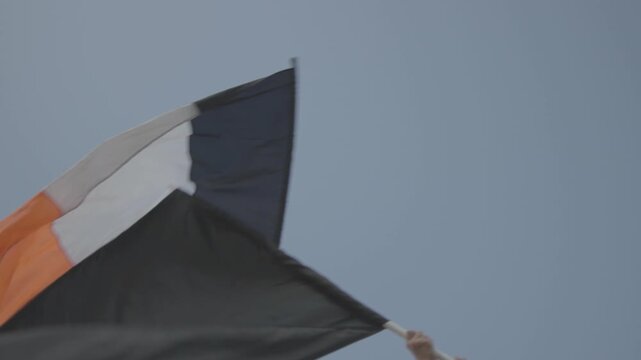 Historical Russian Empire flag with black, yellow, and white stripes waving alongside a solid black banner against a clear blue sky during a windy day outdoors.

