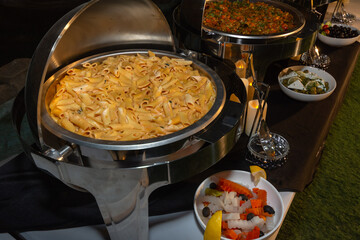A large stainless steel chafing dish filled with creamy penne pasta at a catered buffet.