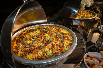 A lavish buffet table illuminated at night, featuring hot biryani rice, various fresh salads, and olives in elegant chafing dishes and bowls.
