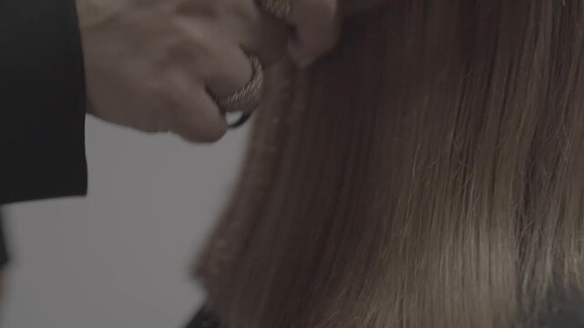 Close-up of professional stylist hands trimming long blonde hair. Hairdresser uses comb and scissors to create a straight blunt cut on a female client in a beauty salon.

