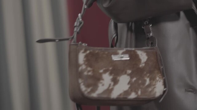 Fashionable woman modeling a long brown leather coat, matching high-heeled boots, and a cow-print handbag. Elegant autumn outfit details showcased in a professional studio setting with red curtains.

