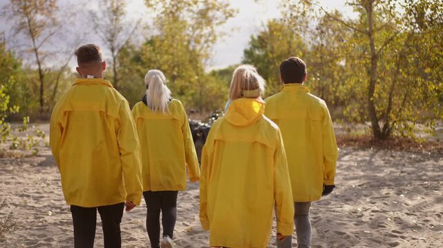 Slow motion. Four white friends in yellow raincoats approaching parked atv on sandy forest trail, autumn trees backlit by sun, group rendezvous with driver, anticipation