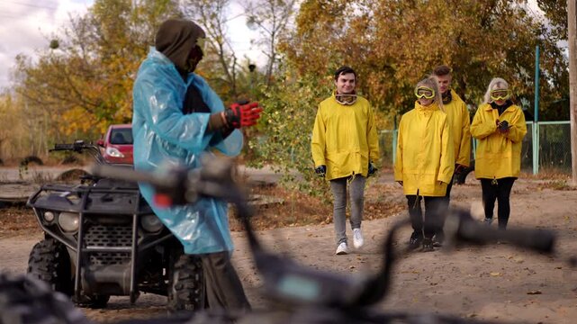 Driver in blue poncho greeting four white friends by ATV with gloved hands and goggles, autumn foliage and mud tracks visible, safety briefing and toolkit