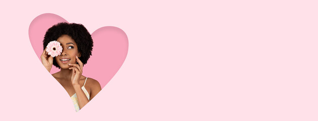 A cheerful woman with curly hair is smiling while holding a flower up to her face. The background is a gentle pink, creating a fun and vibrant atmosphere. The image expresses joy and beauty.