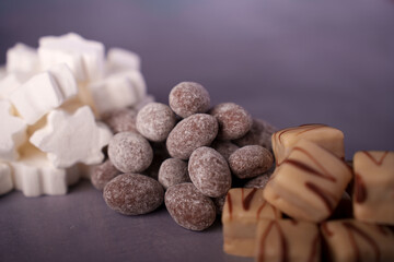 assorted sweets composition featuring white marshmallow stars, chocolate-coated almonds, and caramel cubes on a soft purple surface, selectivefocus, styled, appetizing, pattern, shapes, focus, scene