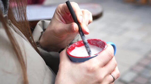 Young woman drinking hot cocoa at a Christmas market. Cozy winter atmosphere, festive lights and decorations in the background. Warm drink with whipped cream in hands, cold weather, holiday mood, slow