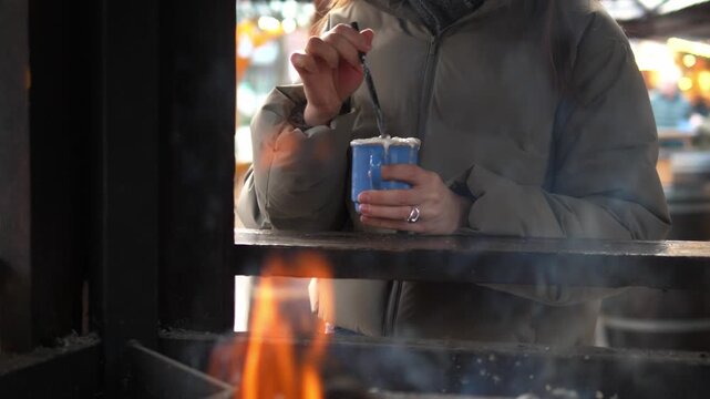 Young woman drinking hot cocoa at a Christmas market. Cozy winter atmosphere, festive lights and decorations in the background. Warm drink with whipped cream in hands, cold weather, holiday mood, slow
