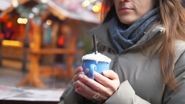 Young woman drinking hot cocoa at a Christmas market. Cozy winter atmosphere, festive lights and decorations in the background. Warm drink with whipped cream in hands, cold weather, holiday mood, slow