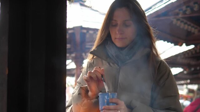 Young woman drinking hot cocoa at a Christmas market. Cozy winter atmosphere, festive lights and decorations in the background. Warm drink with whipped cream in hands, cold weather, holiday mood, slow
