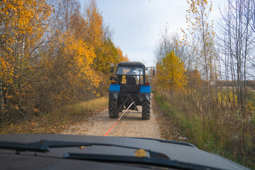 A tractor is towing a broken car on a rural dirt road, an autumn day