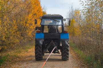 A tractor is towing a broken car on a rural dirt road, an autumn day
