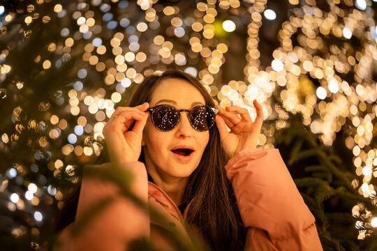A surprised girl wearing sunglasses at a Christmas market in Europe. In the background, bokeh from the lights, with reflections of the market lights in her sunglasses.