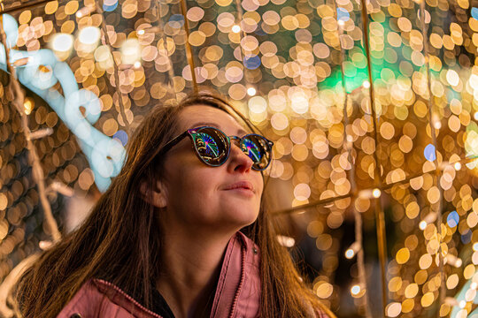 A surprised girl wearing sunglasses at a Christmas market in Europe. In the background, bokeh from the lights, with reflections of the market lights in her sunglasses.