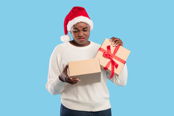 Celebrating the holiday season, a person wearing a Santa hat shows a look of disappointment while holding two gift boxes, one wrapped with a red bow. The background is bright blue.