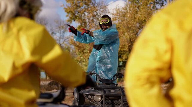 Instructor in blue poncho splashing water toward group of riders in yellow jackets, energetic demonstration at park with autumn trees and sunlit sky, observers